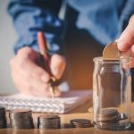 A person's hand inserts a coin into a jar for savings, with a notebook and pen nearby, showcasing financial planning.