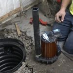 Worker servicing a cylinder-shaped submersible pump in an open sump pit, with tools on a concrete floor.