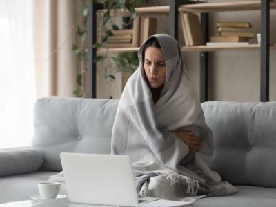 Woman wrapped in a blanket, sitting on a couch with a laptop, looking cold and uncomfortable, illustrating the need for efficient heating in a home.