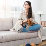 Woman knitting on a couch with yarn and needles in a well-lit living room, featuring a cozy blanket, promoting home comfort.